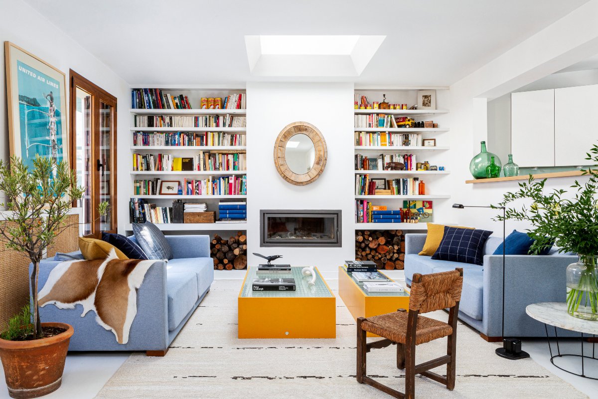 Salón pequeño con dos sofás azules, mesas amarillas y estantería de obra blanca